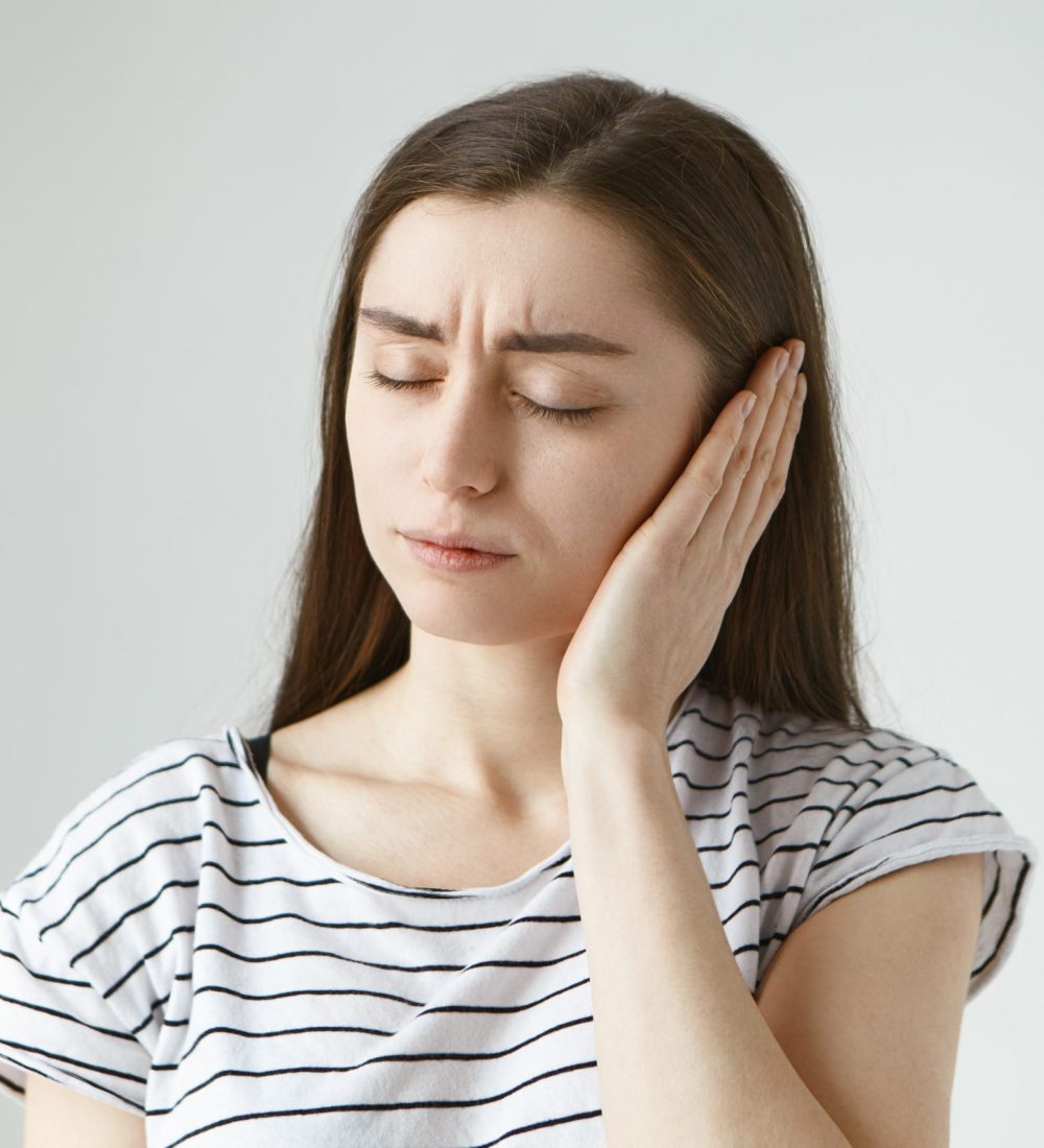 Studio shot of attractive casually dressed young dark haired female keeping eyes closed and covering sore ear with hand while having some problems with hearing. Health, people and pain concept
